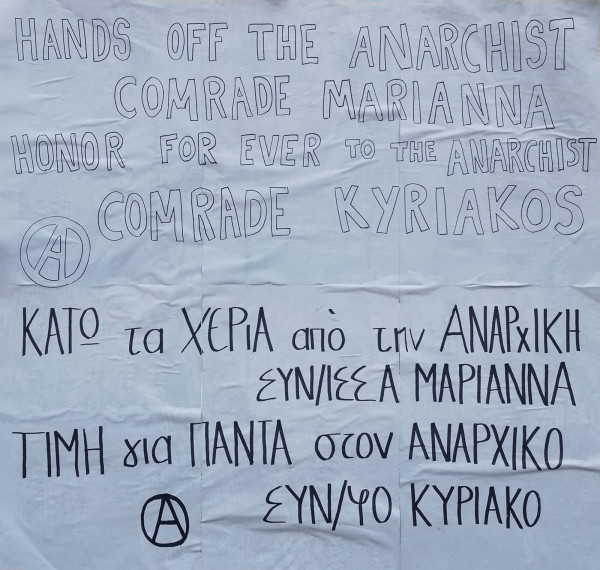 Ein Poster mit hellblauem Hintergrund zeigt handgeschriebene Texte in Schwarz. Oben steht auf Englisch: "Hands off the anarchist comrade Marianna. Honor forever to the anarchist comrade Kyriakos." Darunter folgt derselbe Text auf Griechisch: "Κάτω τα χέρια από την αναρχική συντρόφισσα Μαριάννα. Τιμή για πάντα στον αναρχικό σύντροφο Κυριάκο." Zwei Anarchismus-Symbole, ein eingekreistes A, sind jeweils links vom englischen und griechischen Text platziert.