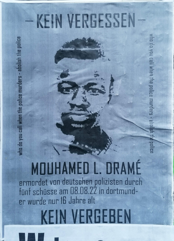 Ein graues Poster zeigt das Porträt von Mouhamed L. Dramé. Oben steht "Kein Vergessen". Seitlich ist in Englisch die Frage "Who do you call when the police murders - abolish the police" zu lesen. Unten steht: "Mouhamed L. Dramé ermordet von deutschen Polizisten durch fünf Schüsse am 08.08.22 in Dortmund - er wurde nur 16 Jahre alt". Am unteren Rand steht "Kein Vergeben". Das Design ist minimalistisch gehalten.