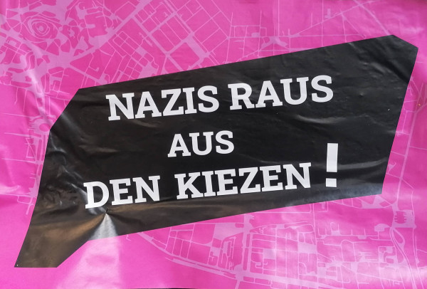 Ein Poster mit einem pinken Hintergrund zeigt eine Stadtkarte. In der Mitte befindet sich eine schwarze, unregelmäßige Form mit weißem Text: "Nazis raus aus den Kiezen!"
